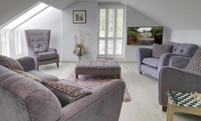Elegant living room with grey armchairs, patterned cushions, and a chess table, bathed in natural light from skylights and tall windows.